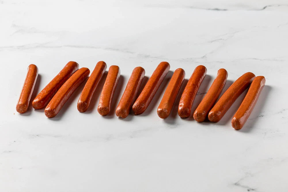 Six uncooked Hot Dogs are arranged in a slightly scattered row on a white marble surface.