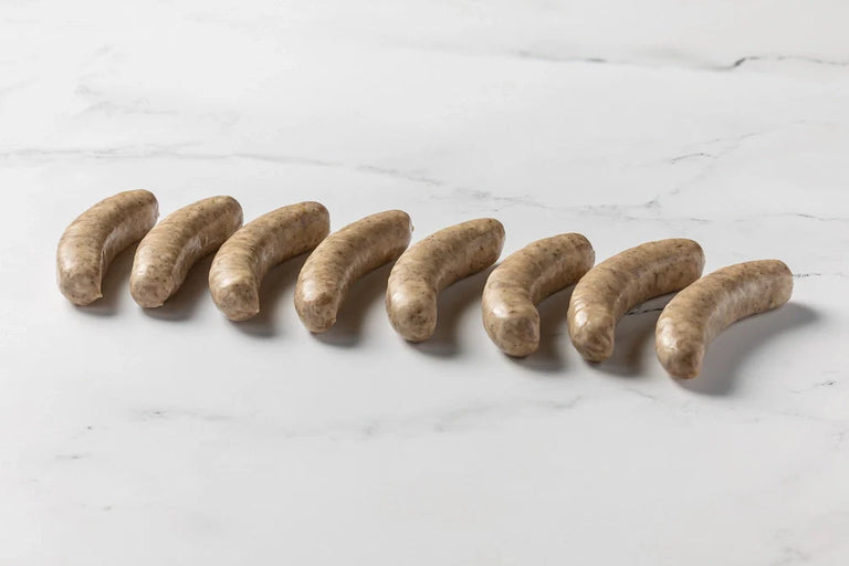 Four uncooked Italian Sausages are lined up on a white surface, sprinkled with black pepper and spices.