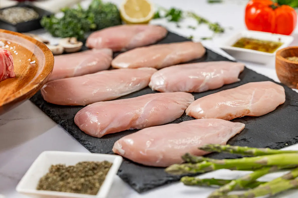 The Essentials Box features 100% American raw chicken breasts displayed on a black slate alongside fresh asparagus, herbs, spices, tomato, and lemon for a vibrant presentation.