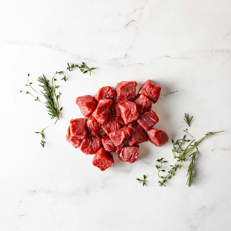 Grass Fed Beef Steak Tips are arranged on a white marble surface, garnished with fresh rosemary and thyme, ready to be packed into convenient packs for your next meal.
